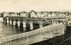 Historische Ansicht der Brücke über die Maas bei Maastricht. Eine hohe Kaimauer schützt die Wohnhäuser vor Hochwasser, rechts Fabrikschornsteine / Schlote von Fabrikanlagen. Im Vordergrund führt eine Rampe zur Straße und Brücke.