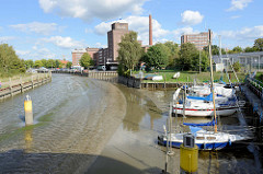 Die Krückau in Elmshorn bei Niedrigwasser / Ebbe;  nur ein schmales Rinnsal zeigt die Fahrrinne an; die Sportboote sind trockengefallen und liegen im Schlick - im Hintergrund die Industriearchitektur der Köllnflockenwerke.
