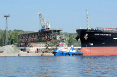 Hafenanlage in Stettin, Kran auf einem Laufgerüst; der Frachter Jose Progress wird von einem FairPlay-Schlepper für die Ausfahrt vorbereitet.