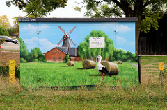 Verteilerkasten einer Energiestation mit Illusionmalerei / Fassadenbild versehen. Weißstorch auf einer Wiese mit Heuballen, dahinter eine Windmühle mit Scheune.