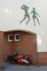 Bronzefiguren, tanzende Frauen als Relief an einer Hausfassade in der Harmsstraße von Hamburg-Harburg; Ein rotes Motorrad ist im Hausdurchgang geparkt.
