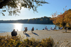 Herbsttag  an der Promenade der   Außenmühle im Stadtpark von Hamburg Harburg;  sonnenhungrige Parkbesucherinnen  sitzen auf Einzelstühlen am See in der Sonne. Die die Blätter von Bäumen und Sträuchern sind herbstlich gefärbt.
