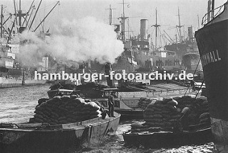 Fotodokumentation - Geschichte der Arbeit im Hamburger Hafen. Geschäftiges Treiben im Hamburger Hafen ca. 1930; Frachter liegen an den Duckdalben in der Mitte des Segelschiffhafens. Ein Schlepper zieht unter Dampf eine Schute im Hafenbecken.