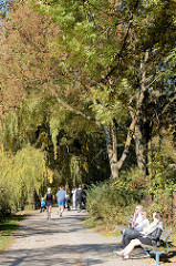 Herbst in der Hansestadt Hamburg, Besucherinnen der Grünanlage am Weiher in Eimsbüttel sitzen in der Sonne auf Parkbänken, joggen,  fahren Fahrrad oder gehen unter den Herbstbäumen spazieren.