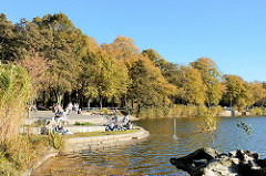 Herbsttag an der Promenade der Außenmühle im Stadtpark von Hamburg Harburg; sonnenhungrige Parkbesucherinnen sitzen auf Einzelstühlen am See in der Sonne. Die die Blätter von Bäumen und Sträuchern sind herbstlich gefärbt.