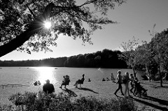 Sonnentag an der Promenade der   Außenmühle im Stadtpark von Hamburg Harburg; Spaziergänger schlendern um den kleinen See andere sitzen auf Einzelstühlen in der Sonne. Gegenlichtsaufnahme, die tief stehende Sonne strahlt durch die Blätter eines Baum