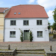 Historisches Wohnhaus, Fachwerkgebäude mit weißer Fassade und Rosenstöcke am Eingang in der Straße frische Grube in Wismar; das Gebäude steht als herausragendes Baudenkmal der Hansestadt unter Denkmalschutz. Im Vordergrund ein Treppenabgang zum Mühle