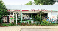 Möbel-Paradies, leerstehender Möbeldiscounter in der Philipp Müller Straße von Wismar; der Eingangsbereich ist mit jungen Bäumen und Wildkraut bewachsen, die Fensterscheiben mit Plakaten beklebt.