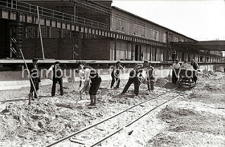 Der Hafenkai am Kaischuppen 58 wird gebaut; Bauarbeiter schaufeln Sand - drei Männer schieben auf provisorischen Gleisen eine mit Sand gefüllte Lore der Laderampe entlang.