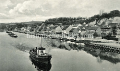 Schlepperverband mit mehreren Lastkähnen und einem Dampfschlepper mit hoher Schornstein auf der Alten Oder vor Oderberg. Kleine Boote liegen am Ufer, auf einer Wiese am Wasser wird Wäsche getrocknet.