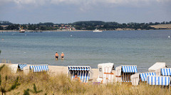 Strand mit Strandkörben an der Ostsee im Ostseebad Boltenhagen; zwei Touristen gehen im seichten Ostseewasser - im Hintergrund die Ostseeküste bei Redewisch.