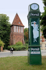 Alte Normaluhr mit der Reklame für das Waschmittel Persil; weiße Frau mit Hut hält eine Packung;  im Hintergrund der historische Wasserturm / Stadttor, der ein Teil der historischen Wehranlage von Wismar ist.