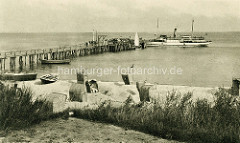 Altes Foto von der Seebrücke in Kühlungsborn, die 1929 errichtet wurde. An dem Landungssteg legten Dampfschiffe/Ausflugsschiffe an. Im Vordergrund der Strand mit Sandburgen und Strandkörben, einige  Ruderboote sind an Land gezogen.