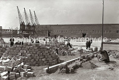 Der Platz vor dem Verteilungsschuppen 58 am Togokai des Südwesthafens wird von Straßenarbeitern gepflastert. Die Laderampen sind für die Be- und Entladung von LKW vorgesehen. Eine Schubkarre aus Holz liegt auf der Seite.