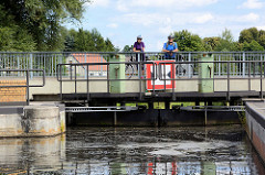 Geschlossenes Schleusentor der Schleuse Schöpfurth am Finowkanal; die Schleusenkammer hat eine Länge von 41m und einen Hub von 3,60m. Auf der Fussgängerbrücke stehen FahrradfahrerInnen und sehen der Schleusung zu.