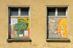 Vernagelte Fenster eines leer stehenden Gebäudes in Güstrow, die Holzplatten sind mit bunter Graffiti- Porträts versehen.
