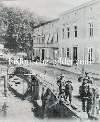 Historische Ansicht vom  Finowkanal bei Eberswalde; Fischer stehen am Ufer an dem Ketscher trocknen. Eine Waage mit einem Eimer zum Abwiegen von Fischen steht am Weg, im Hintergrund sitzt eine alte Frau auf einer Holzbank. Am Kai ist ein Holzbo