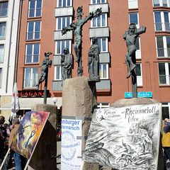 Ostermarsch 2019 - Demo für Abrüstung in Hamburg. politische Plakate vor den Bronzeskulpturen der Kreuzigungsgruppe vor der  Dreieinigkeitskirche von Hamburg St. Georg.