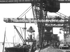 Die Kohleladung eines Frachtschiffs wird am Kai gelöscht. Krananlagen holen die Fracht aus dem Laderaum und bringen die Kohle an Land - ein Lastwagen mit Anhänger steht unter einer Schütte und wird beladen.