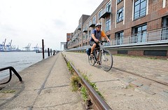 Schienen des Hafenkrans am Kai vor dem Altonaer Kaispeicher. Ein Fahrradfahrer fährt auf dem Kopfsteinpflaster die Kaianlage entlang.