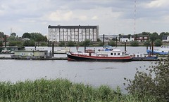 Blick über den Holzhafen in Hamburg Moorfleet - auf der gegenüber liegendes Seite der Moorfleeter Deich und die typische Industriearchitektur eines Lagergebäudes. An den Bootsstegen des ehemaligen Laufs der alten Elbe sind Boote vertäut.