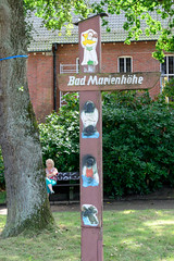 Fotos aus dem Hamburger Stadtteil Sülldorf im Bezirk Hamburg Altona. Hölzernes Hinweisschild für das Bad Marienhöhe - Schnitzereien sind mit Farbe verschmiert.