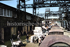Historische Aufnahme vom Hamburger Hafen; unter Portalkränen stehen Eisenbahnwaggons, die mit Ballen beladen werden.